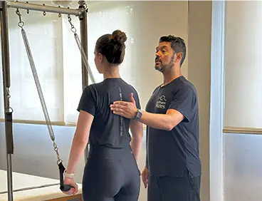 moça se alongando em equipamento de pilates ao lado de um instrutor