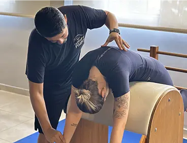 moça se alongando em equipamento de pilates ao lado de um instrutor